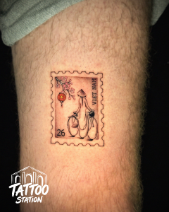 Fine Line Tattoo of a vintage Vietnam postage stamp featuring a woman in ao dai, cherry blossoms and Hoi An lantern by Tattoo Station Hoi An